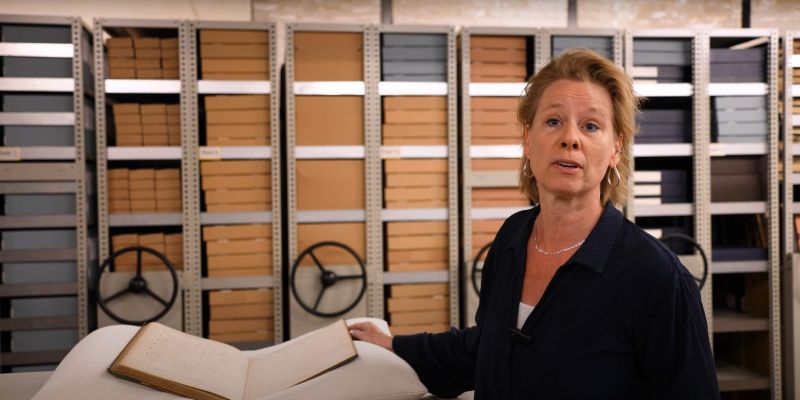 Bezoekster leest een oud scheepsjournaal in een vitrine tijdens de expositie Opgedoken in het Maritiem Museum Rotterdam, met historisch handschrift en kaft zichtbaar.