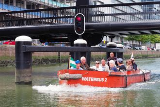 Een Waterbus vaart over de rivier met het Maritiem Museum Rotterdam op de achtergrond, vastgelegd door Theo de Man op een heldere dag met strakblauwe lucht.