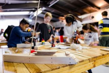 Close-up van kinderen en gereedschap op een werkbank terwijl ze samen een houten scheepsmodel bouwen tijdens de activiteit Schepenbouwers in het Maritiem Museum Rotterdam.